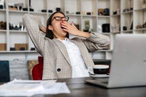Beautiful sleepy woman in eyeglasses yawning while working with laptop and papers indoors