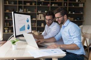 Two businessmen brothers twins working on analytics using computer, sit at desk, review sales statistics charts, graphs shown on pc monitor, discussing project revenue. Family business, partnership